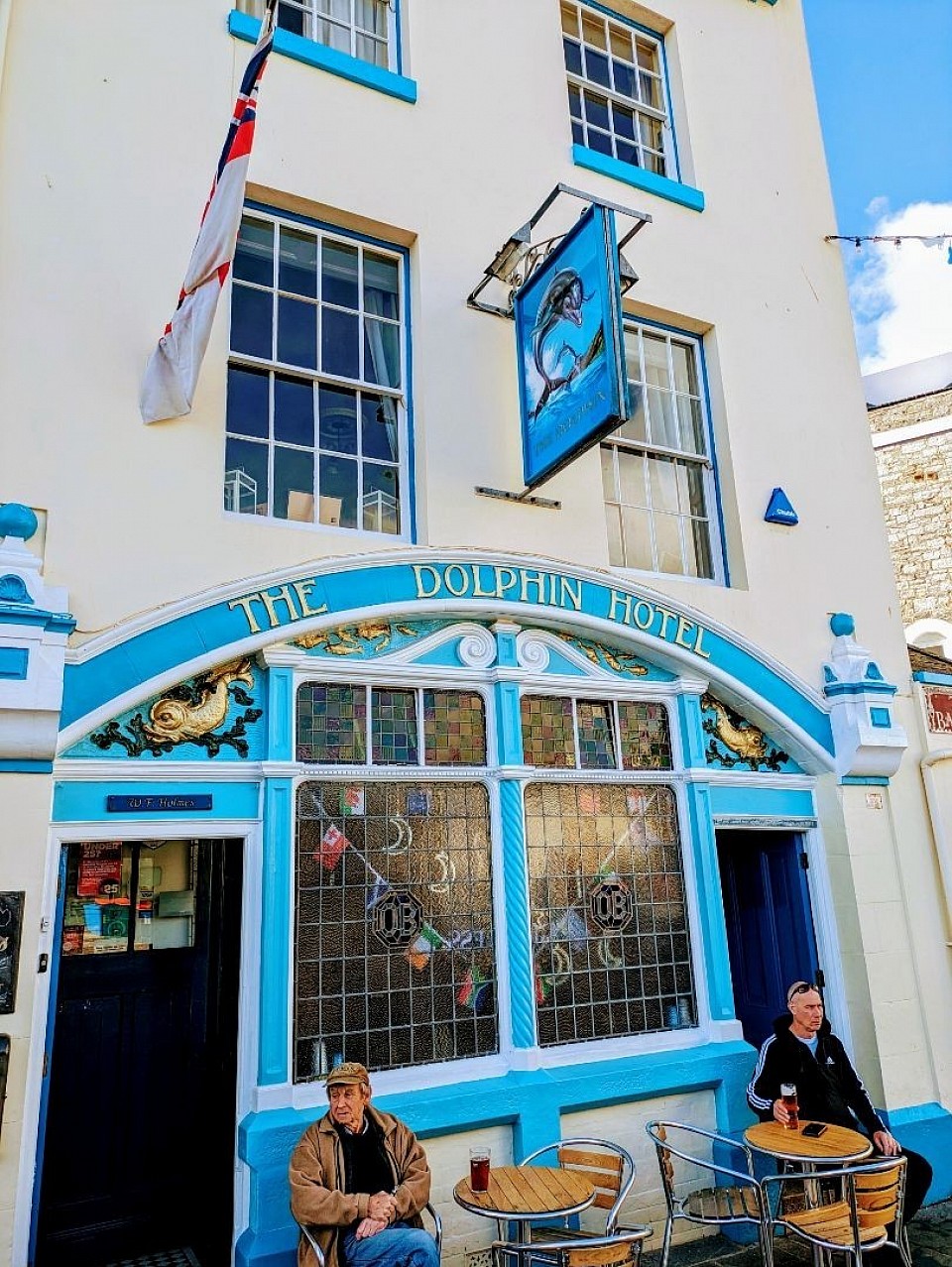 The blue and white exterior of the historic Dolphin Hotel with two men sitting at outdoor tables, lost in contemplation under a sunny sky.
