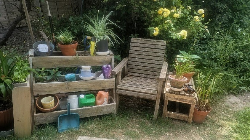A quiet, rustic garden seating and potting corner featuring a wooden slatted armchair, a small side table with potted grasses, and a three-tiered wooden storage shelf organized with terracotta pots, young plants, and gardening supplies against a lush green background.